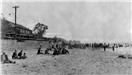 Black and white photo of people seated on the beach with cars and bath house beyond