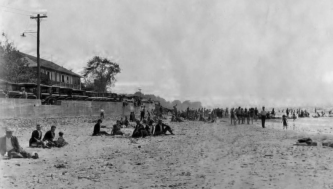 Black and white photo of people seated on the beach with cars and bath house beyond